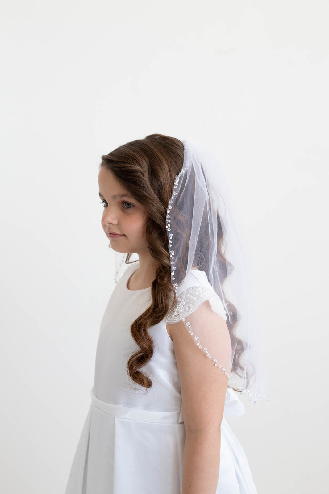 A young girl with long, curly hair wearing a white dress and a veil stands against a plain white background.
