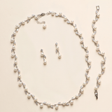 A necklace and earrings set made of white pearls and silver leaf-shaped charms are displayed on a light-colored background.
