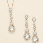 A gold necklace with a pendant and a pair of gold earrings with a pendant are displayed against a light beige background.