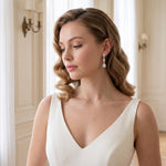 A woman in a white dress with a pensive expression, wearing diamond earrings, stands in a room with white walls and a window.