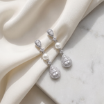 A pair of elegant diamond earrings with pearls and sparkling stones are displayed on a white fabric surface.