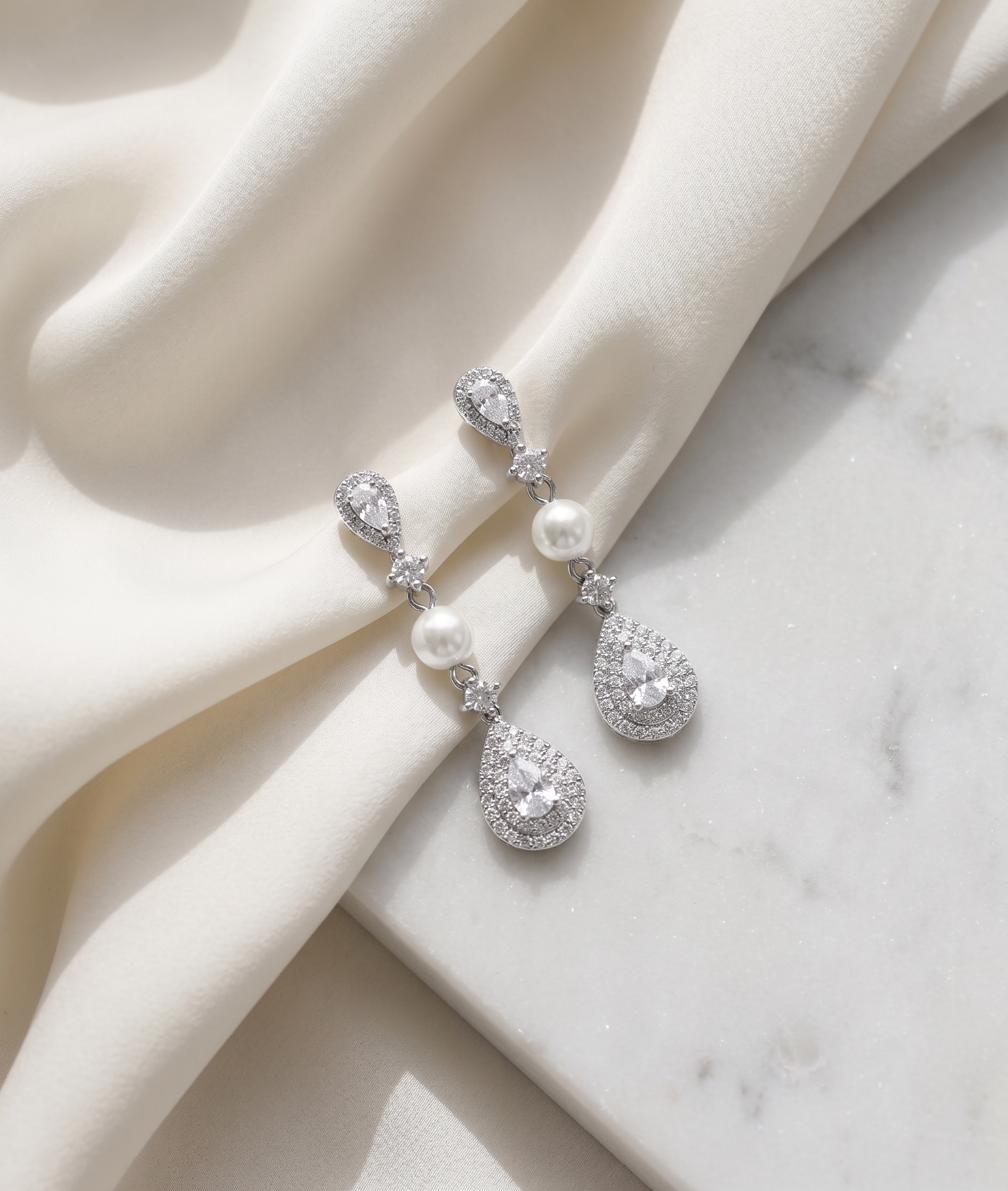 A pair of elegant diamond earrings with pearls and sparkling stones are displayed on a white fabric surface.