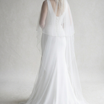 A woman in a long white wedding dress with a veil stands against a plain white background.