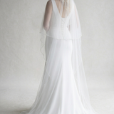A woman in a long white wedding dress with a veil stands against a plain white background.