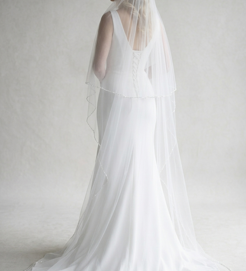 A woman in a long white wedding dress with a veil stands against a plain white background.
