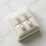 A pair of elegant earrings with pearls and diamonds displayed in a white jewelry box on a marble surface.