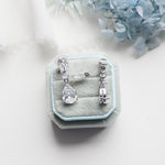 A pair of elegant diamond earrings is displayed in a light blue velvet box, with a white lace cloth and blue flowers in the background.