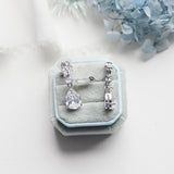 A pair of elegant diamond earrings is displayed in a light blue velvet box, with a white lace cloth and blue flowers in the background.