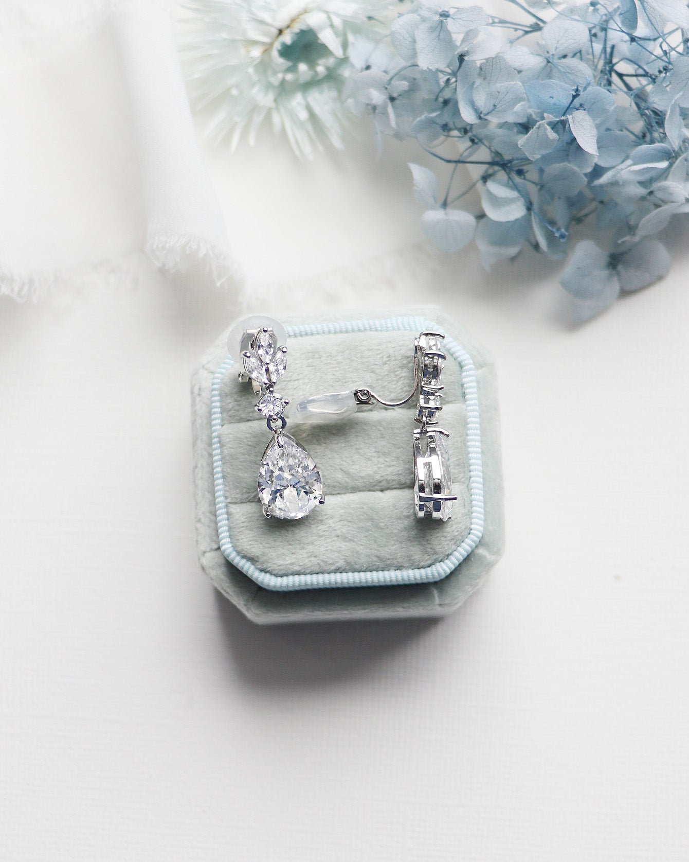 A pair of elegant diamond earrings is displayed in a light blue velvet box, with a white lace cloth and blue flowers in the background.