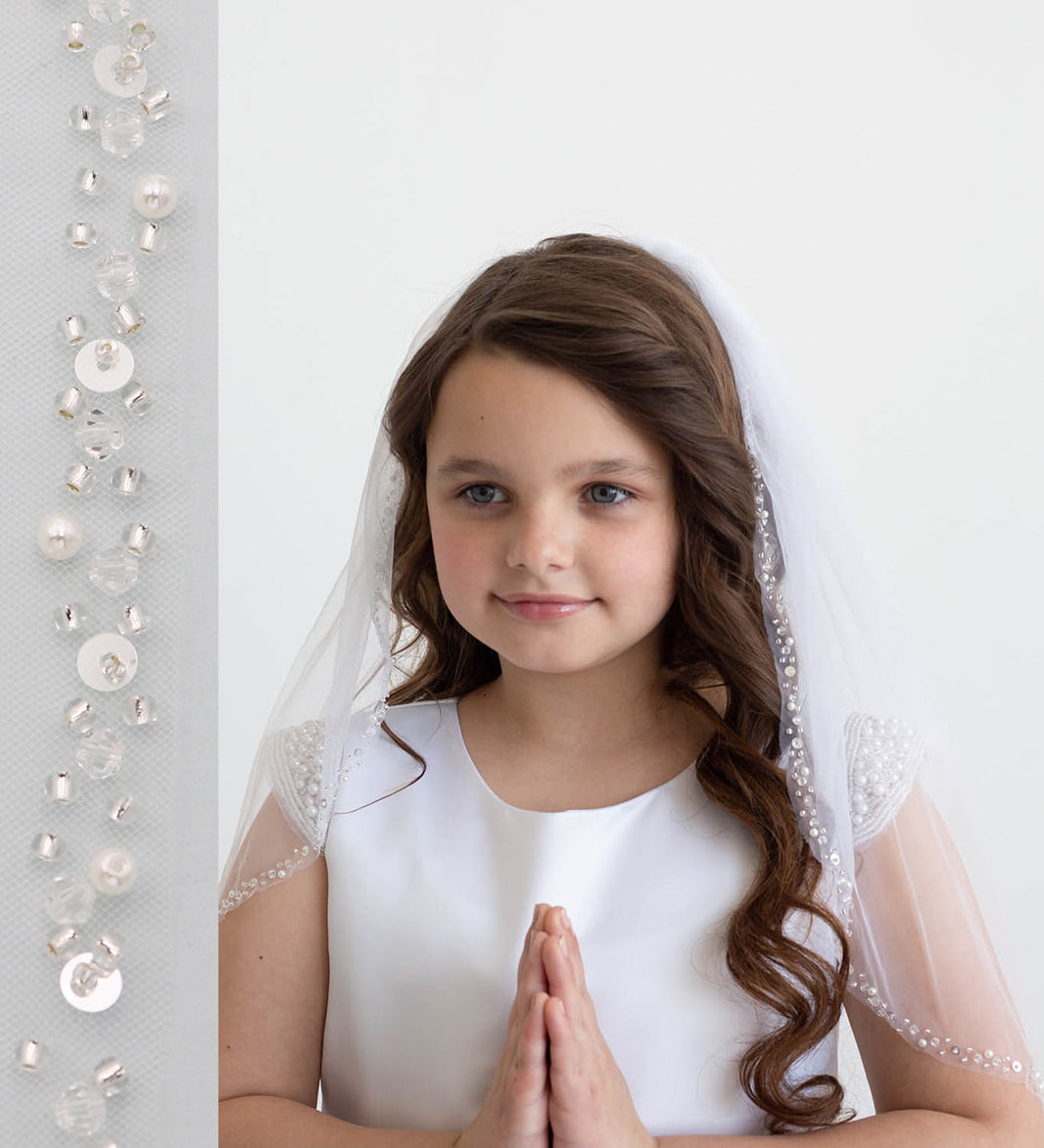 A young girl with long brown hair wearing a white dress and veil stands in front of a white background, smiling at the camera.