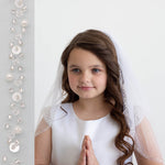 A young girl with long brown hair wearing a white dress and veil stands in front of a white background, smiling at the camera.