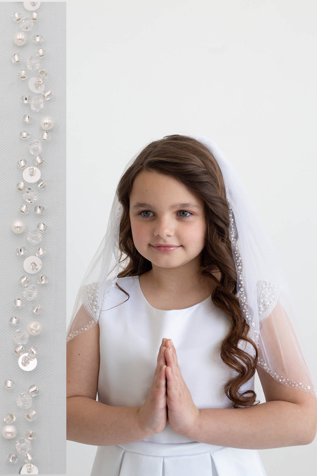 A young girl with long brown hair wearing a white dress and veil stands in front of a white background, smiling at the camera.