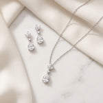 A necklace and earrings are displayed on a white marble surface, with a delicate fabric draped over the background.