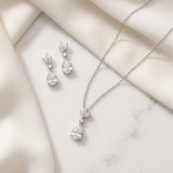 A necklace and earrings are displayed on a white marble surface, with a delicate fabric draped over the background.