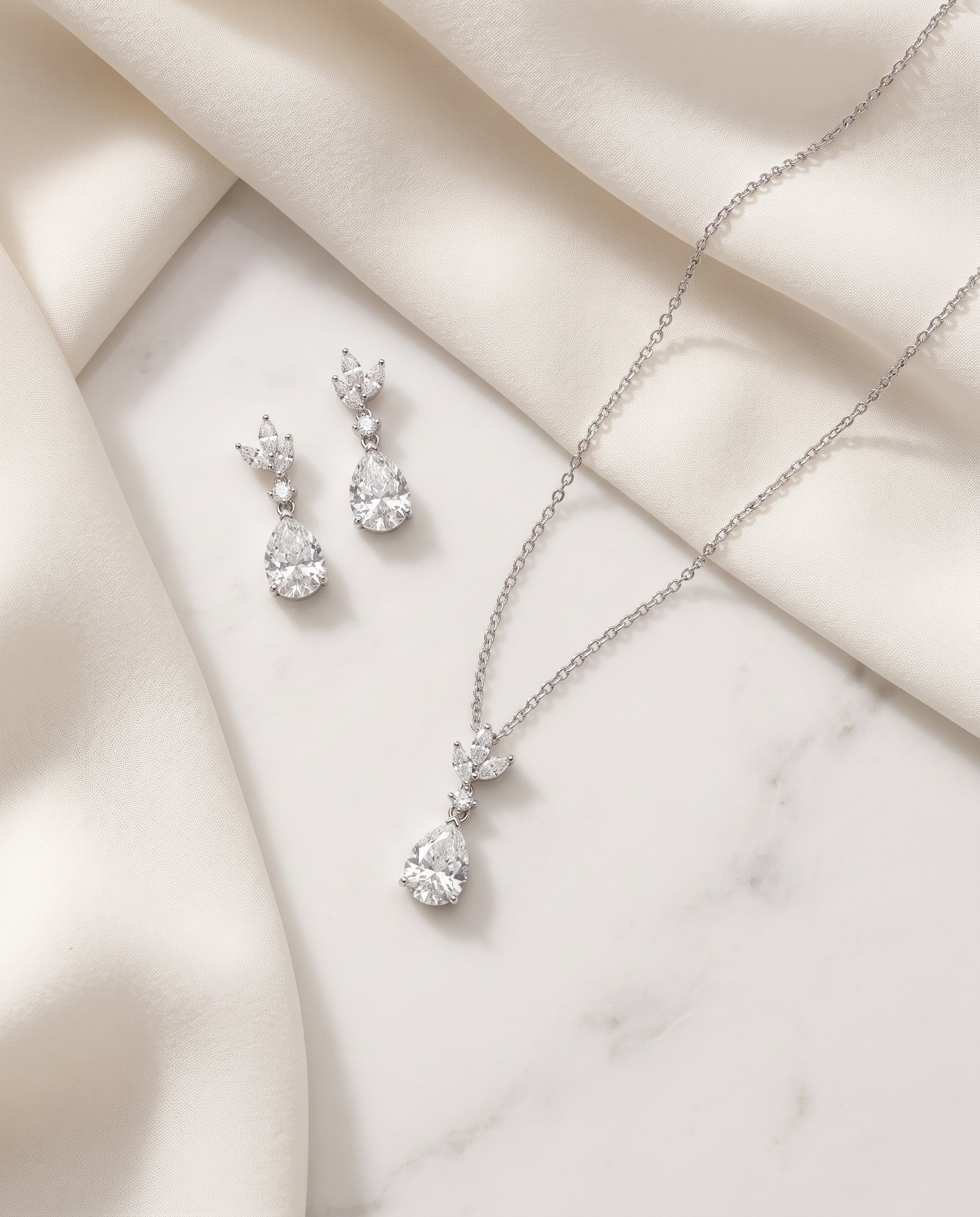 A necklace and earrings are displayed on a white marble surface, with a delicate fabric draped over the background.