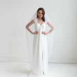 A woman in a white wedding dress stands with her hands on her hips, wearing a long white veil that extends behind her.