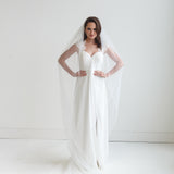 A woman in a white wedding dress stands with her hands on her hips, wearing a long white veil that extends behind her.