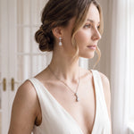 A woman wearing a white dress and earrings stands in front of a window, gazing off to the side.