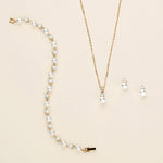 A delicate gold necklace with a pendant and earrings are displayed on a light beige background.