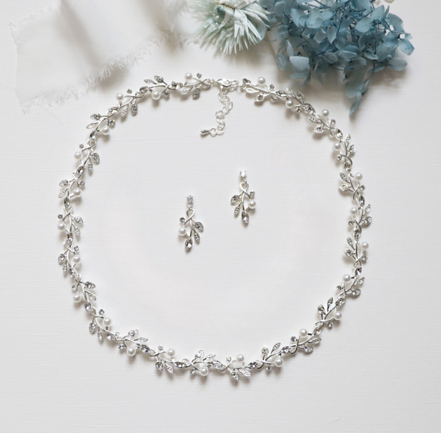 A delicate necklace with a leaf-shaped pendant, earrings, and a bracelet, all adorned with small white pearls and silver leaves, arranged on a white surface.