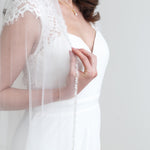 A woman in a white wedding dress with a long veil stands against a white background.