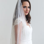 A woman in a white wedding dress with a long veil stands against a plain white background, gazing off to the side.