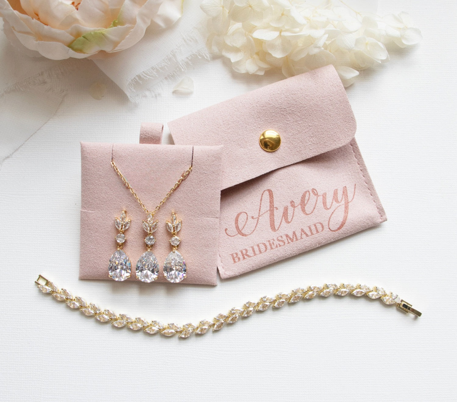 A bridesmaid's jewelry set, including a necklace with three dangling crystal pendants and a bracelet, is displayed on a white surface alongside a small pink jewelry box and a white flower.