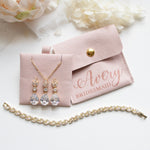 A bridesmaid's jewelry set, including a necklace with three dangling crystal pendants and a bracelet, is displayed on a white surface alongside a small pink jewelry box and a white flower.