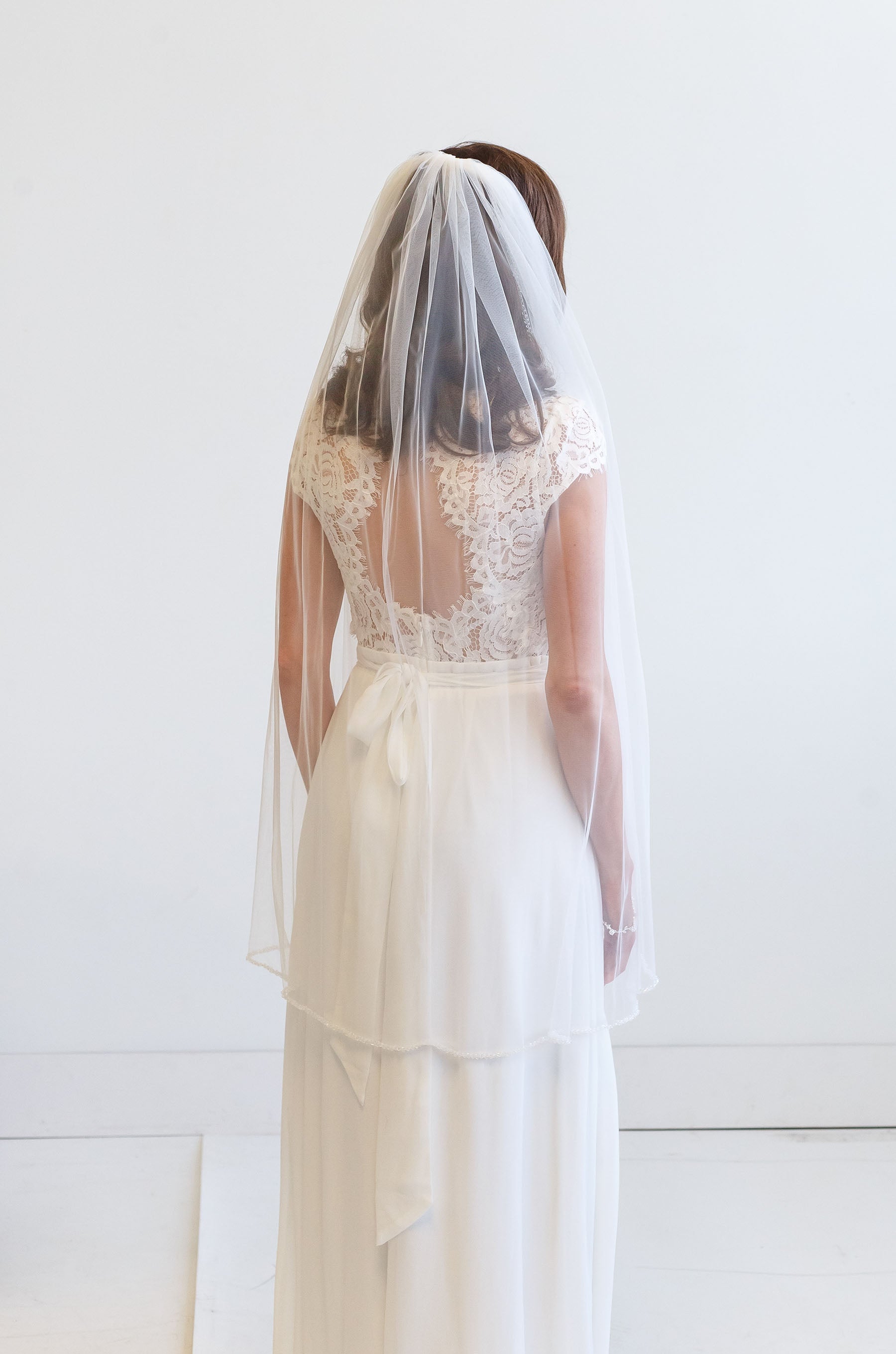 A woman wearing a white wedding dress with a veil, standing against a white background.