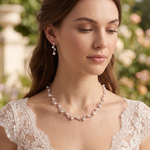 A woman wearing a white lace dress and a pearl necklace stands in front of a garden with greenery in the background.