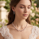 A woman wearing a white lace dress and a pearl necklace stands in front of a garden with greenery in the background.