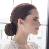 A woman with a bun hairstyle wearing a white lace top and large dangling earrings, looking to the side with a neutral expression.