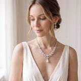 A woman wearing a white dress and a necklace with a pendant stands in front of a window.