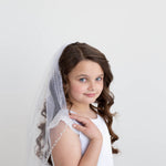 A young girl with curly brown hair wearing a white dress and veil stands against a plain white background, smiling at the camera.