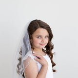 A young girl with curly brown hair wearing a white dress and veil stands against a plain white background, smiling at the camera.