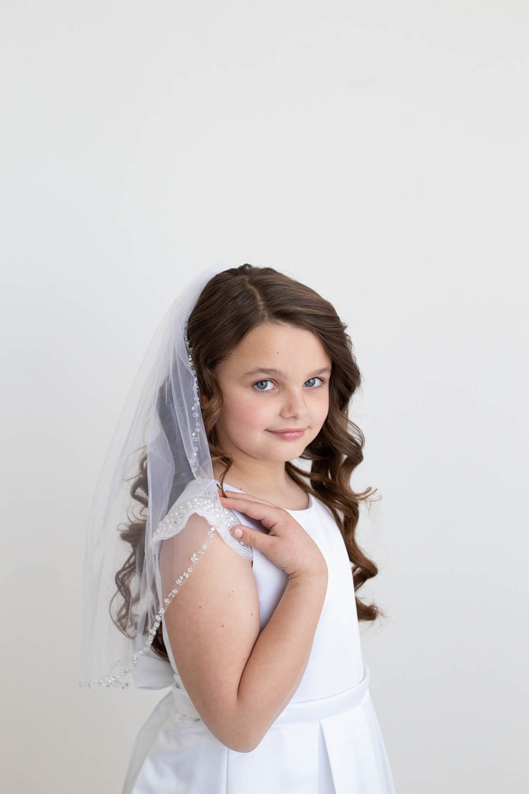 A young girl with curly brown hair wearing a white dress and veil stands against a plain white background, smiling at the camera.