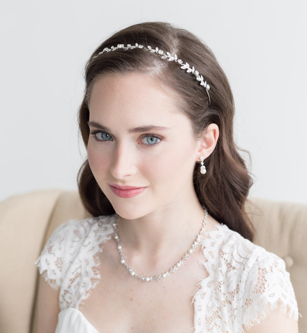A woman with long brown hair wearing a white lace dress and a headband with small white flowers, smiling at the camera.