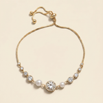 A gold-colored bracelet with a central pearl and a large crystal accent, set against a light beige background.