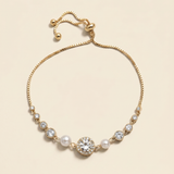 A gold-colored bracelet with a central pearl and a large crystal accent, set against a light beige background.