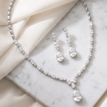A necklace and earrings with large, clear gemstones are displayed on a marble surface, with a beige fabric draped over the background.