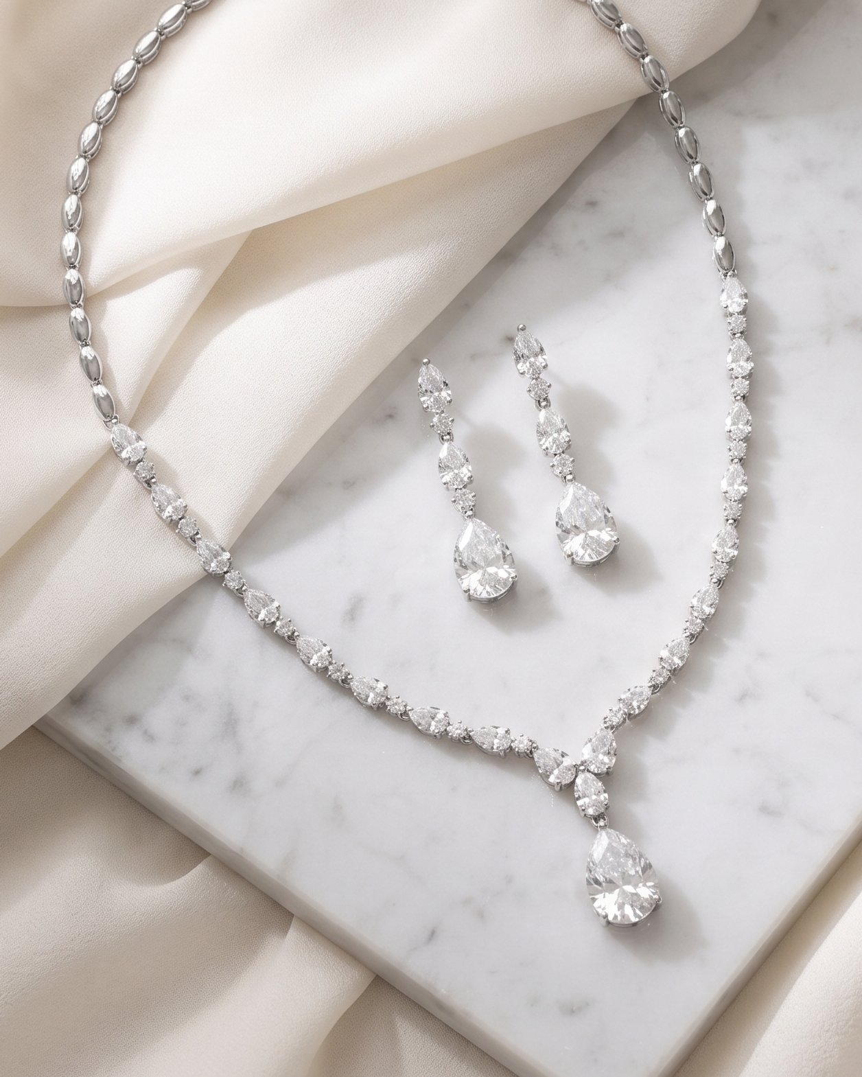 A necklace and earrings with large, clear gemstones are displayed on a marble surface, with a beige fabric draped over the background.