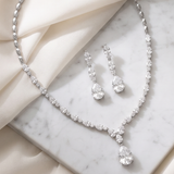 A necklace and earrings with large, clear gemstones are displayed on a marble surface, with a beige fabric draped over the background.