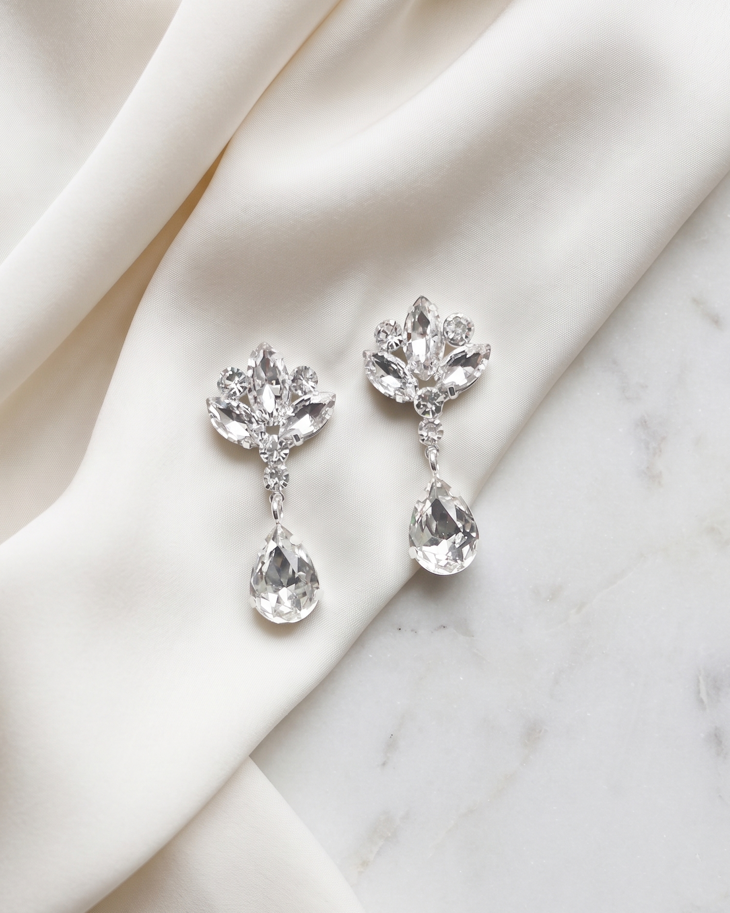 A pair of elegant diamond earrings with clear gemstones, resting on a white fabric surface.