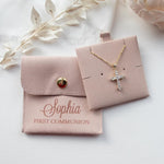 A necklace with a cross pendant and a small pouch with the name "Sophia" printed on it, along with some white flowers and a white fabric background.