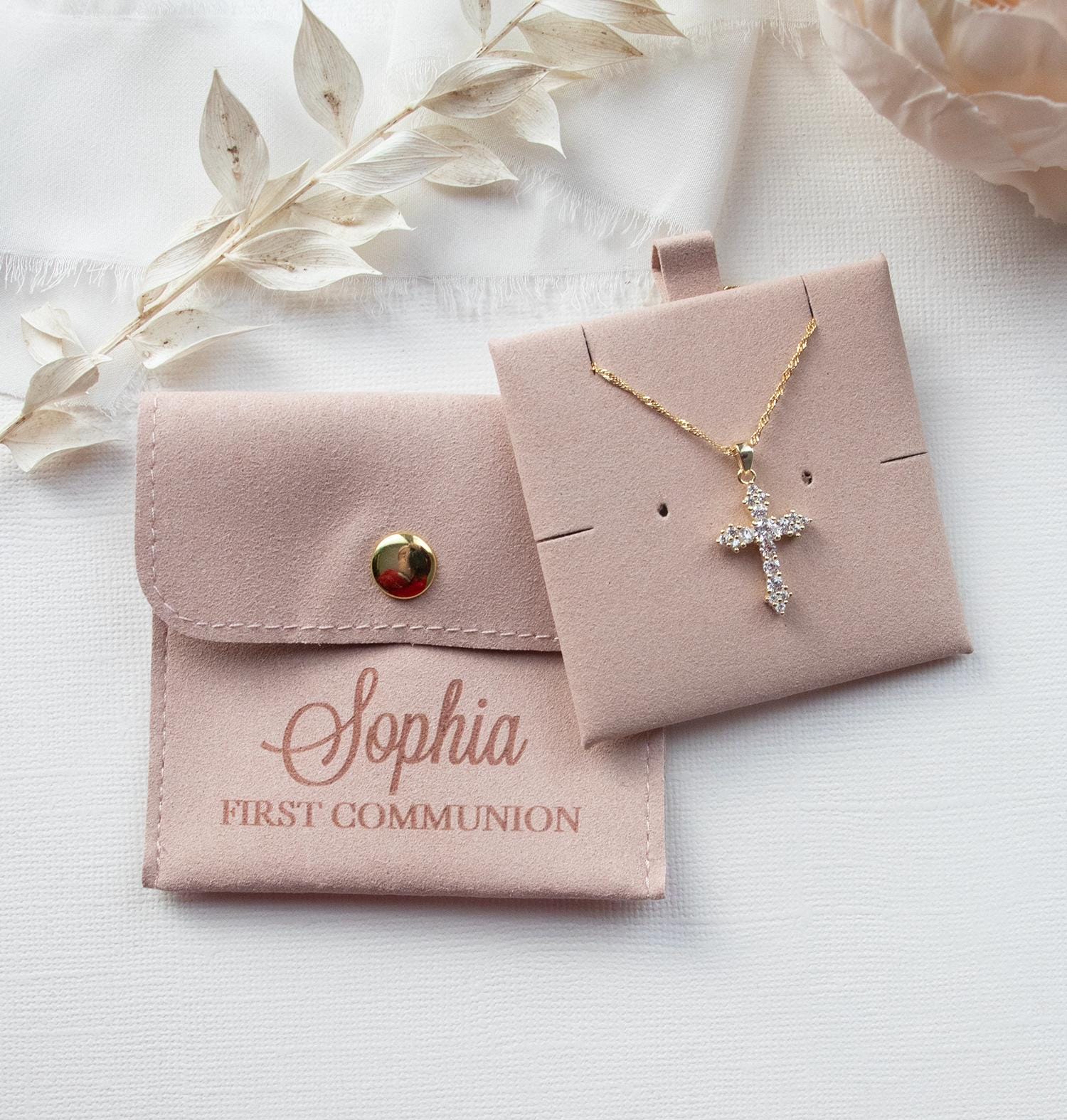 A necklace with a cross pendant and a small pouch with the name "Sophia" printed on it, along with some white flowers and a white fabric background.