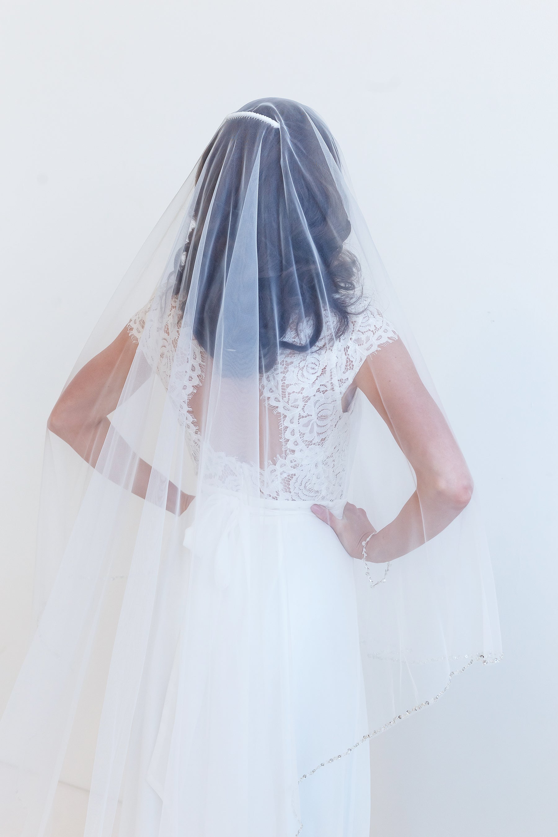 A woman wearing a white wedding dress with a long veil, standing against a plain white background.