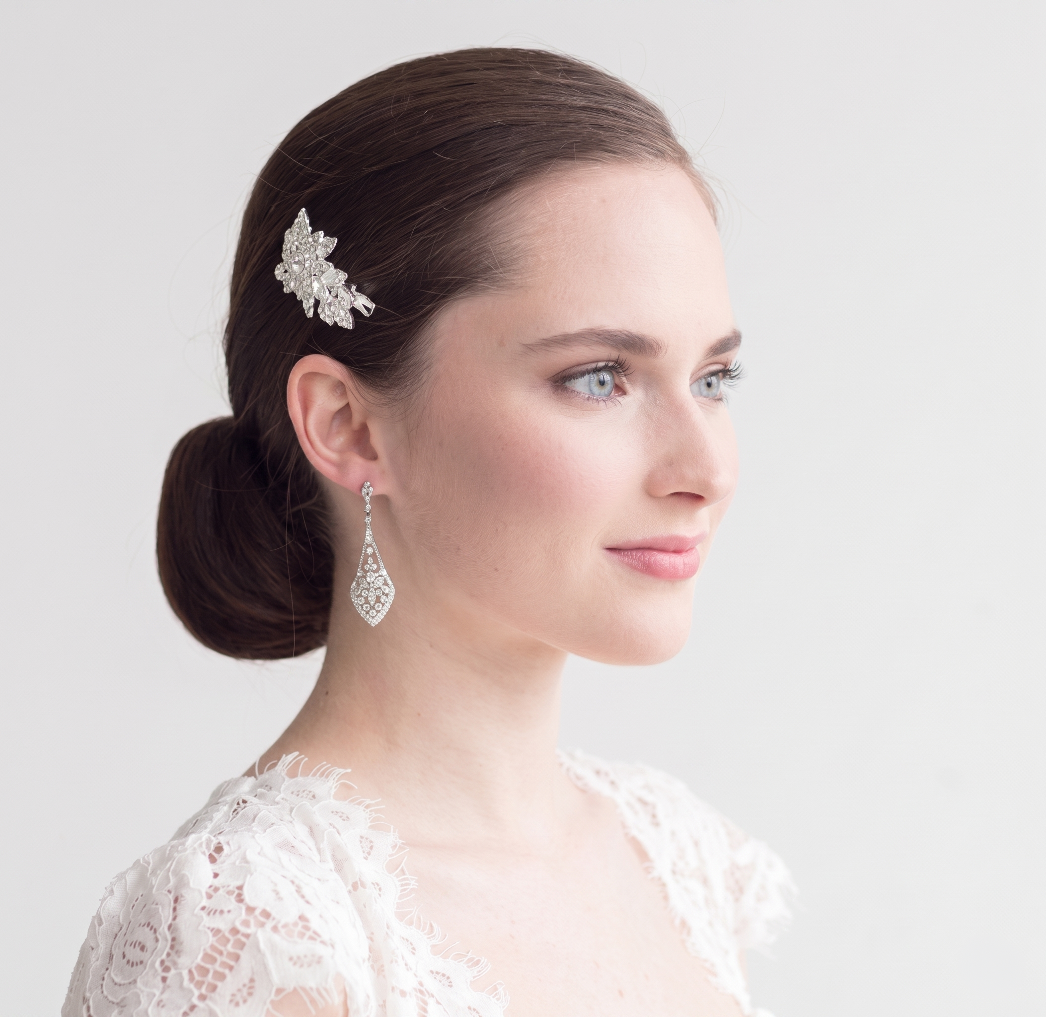 A woman with dark hair styled in a bun, wearing a white lace top and a diamond earring, looking off to the side with a thoughtful expression.