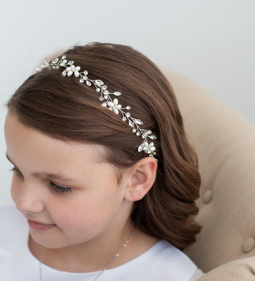 May include: A silver-toned floral headband with pearl and crystal accents. The delicate design features small flowers and leaves, creating an elegant accessory. The headband is worn by a person with brown hair.