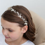 May include: A silver-toned floral headband with pearl and crystal accents. The delicate design features small flowers and leaves, creating an elegant accessory. The headband is worn by a person with brown hair.