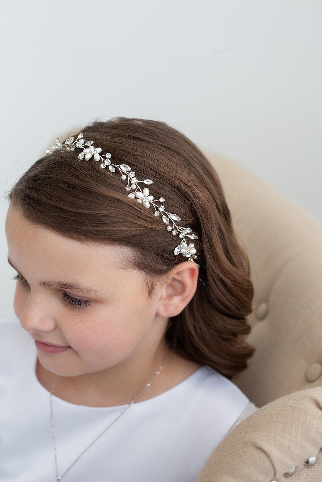 May include: A silver-toned floral headband with pearl and crystal accents. The delicate design features small flowers and leaves, creating an elegant accessory. The headband is worn by a person with brown hair.